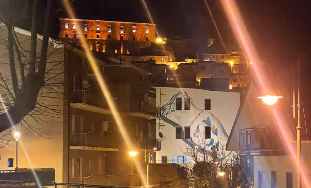 Di notte il Castello illuminato domina il borgo; più in basso il Municipio con la brocca dipinta