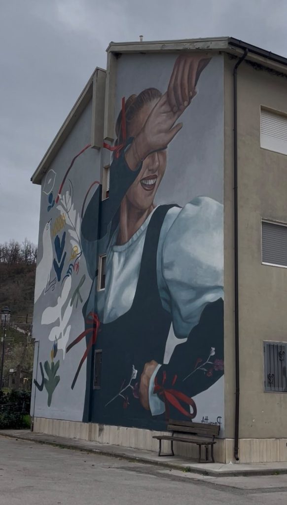 Murale con una giovane in abito tradizionale che danza, con nastri rossi e motivi della ceramica calvellese